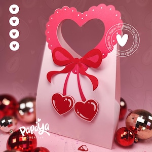 Könnte beinhalten: Eine rosa Geschenkverpackung mit herzförmigem Griff und roter Schleife. Zwei rote herzförmige Kirschen hängen unter der Schleife. Die Tasche ist mit einem wellenförmigen Herzen verziert und von roten und silbernen Weihnachtskugeln umgeben.