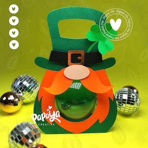Puede incluir: Una caja de regalo con temática de duende verde y naranja con asa, cinturón negro y un trébol. La caja tiene una ventana redonda y transparente. El texto "Papaya Creativa" es visible. Varias bolas de discoteca están esparcidas alrededor de la caja.