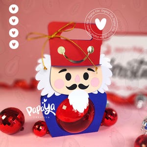 Puede incluir: Una caja de regalo de cascanueces hecha de papel en rojo, azul y blanco, con un asa dorada y un adorno redondo rojo transparente. La caja presenta una cara de dibujos animados y está rodeada de adornos navideños rojos. El logotipo "Papaya Creativa" es visible.