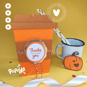 Puede incluir: Una caja de regalo de papel con forma de taza de café en naranja y marrón con una etiqueta dorada festoneada que dice "Thanks for being you". La caja está atada con cordel naranja, negro y blanco junto a una taza de esmalte blanca.