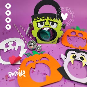 May include: A paper craft set featuring a green Frankenstein monster treat box with a clear circular window, alongside flat paper cutouts of a white ghost, an orange pumpkin, and a vampire face, all arranged with two small disco ball ornaments.