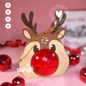 May include: A reindeer-shaped gift box with a red, transparent ball as the nose. The box is tan with brown antlers and a gold ribbon. The text "Papaya Creativa" is printed on the box. Red and silver ornaments are scattered around the box.