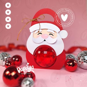 Puede incluir: Una caja de regalo roja con forma de Papá Noel con una ventana redonda y transparente. La caja tiene barba y borde de sombrero blancos, con un asa de cinta dorada. La imagen incluye adornos navideños rojos y plateados y el texto "Papaya Creativa".