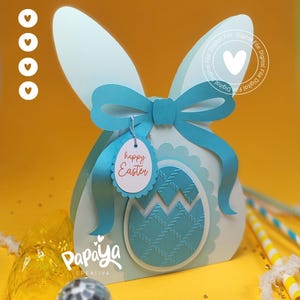 Puede incluir: Una caja de regalo con forma de conejito de Pascua azul claro con un lazo y un diseño de huevo. La caja tiene una etiqueta que dice "Felices Pascuas". El fondo es amarillo.