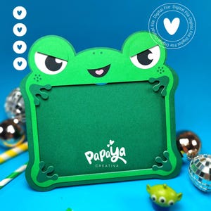 Puede incluir: Un marco de fotos con forma de rana verde que presenta una cara de dibujos animados con ojos grandes y una boca pequeña. El marco tiene cuatro patas de rana en las esquinas y muestra el texto Papaya Creativa en blanco. Un logo digital es visible.