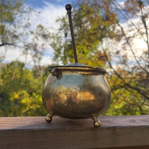 Puede incluir: Un pequeño caldero antiguo de latón con tres patas de bola y una tapa. Un mango largo y delgado de latón se extiende desde la tapa, rematado con un pomo redondo y oscuro. El caldero está sobre una superficie de madera.