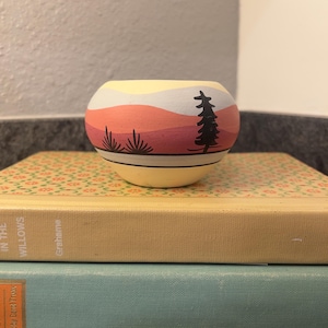 Może przedstawiać: Mały, okrągły, ręcznie malowany ceramiczny garnek z krajobrazem. Projekt przedstawia scenę zachodu słońca z warstwami pomarańczowego, różowego i żółtego, czarne drzewo i stylizowane rośliny. Garnek stoi na dwóch książkach.