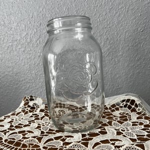 May include: A clear glass mason jar with a rose design. The jar is on a white lace doily. The background is a textured grey wall.