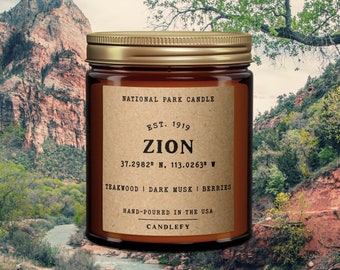 May include: A brown glass candle jar with a gold lid. The label reads "National Park Candle", "Est. 1919", "Zion", "37.2982° N, 113.0263° W", "Teakwood | Dark Musk | Berries", "Hand-poured in the USA", and "Candlefy".