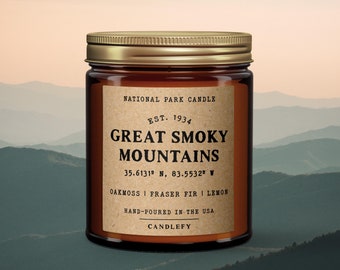May include: A brown glass candle jar with a gold lid. The label reads "National Park Candle Est. 1934 Great Smoky Mountains 35.6131° N, 83.5532° W Oakmoss | Fraser Fir | Lemon Hand-Poured in the USA Candlefy".