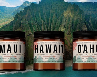 May include: Three brown glass candle jars with black lids and white text labels. The labels read "MAUI", "HAWAII", and "O'AHU" with coordinates and scent descriptions. The candles are set against a backdrop of a lush green mountain range.