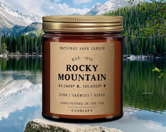 May include: A brown glass candle jar with a gold lid. The label reads "National Park Candle", "Est. 1915", "Rocky Mountain", "40.3428° N, 105.6836° W", "Pine | Oakmoss | Birch", "Hand-poured in the USA", and "Candlefy".