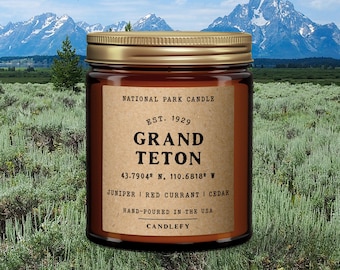 May include: A brown glass candle jar with a gold lid. The label reads "National Park Candle", "Est. 1929", "Grand Teton", "43.7904° N, 110.6818° W", "Juniper | Red Currant | Cedar", "Hand-poured in the USA", and "Candlefy".