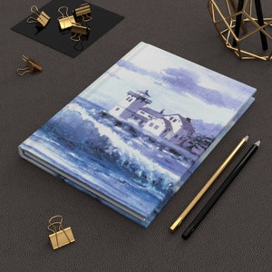 May include: Hardcover journal featuring a watercolor painting of a lighthouse amidst crashing waves in shades of blue and white. Includes gold and black pencils, and gold binder clips. A stylish notebook for writing and sketching.