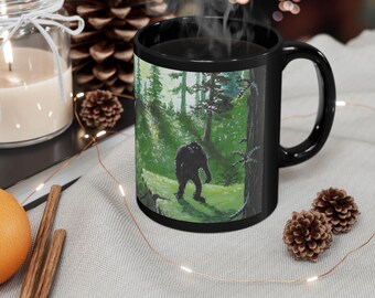 May include: A black ceramic mug featuring a printed illustration of a dark, hairy creature standing in a lush green forest with tall trees. The mug has a curved handle and is filled with a hot beverage emitting visible steam.
