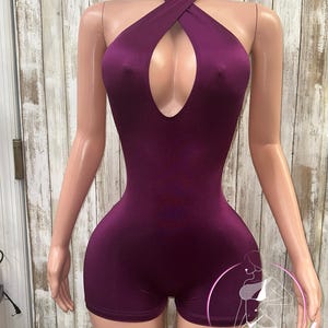 May include: A purple halter-style bodysuit with a criss-cross neckline and a keyhole cutout. The bodysuit has a fitted silhouette and short legs.
