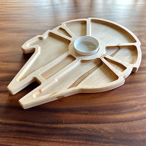 May include: A wooden serving tray shaped like the Millennium Falcon from Star Wars. The tray has multiple compartments and a small, round bowl in the center.