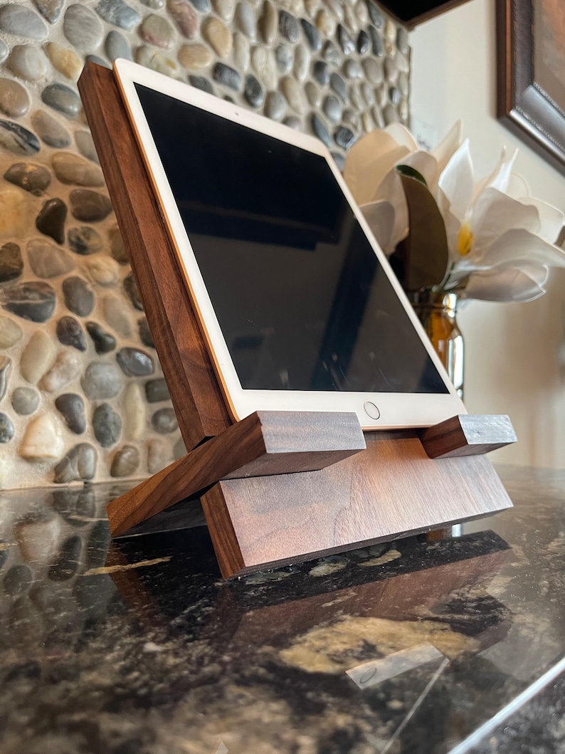 May include: A wooden tablet stand with a dark brown finish. The stand is angled to hold a tablet in an upright position. The stand is sitting on a black countertop.