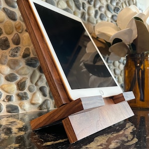 May include: A wooden tablet stand with a dark walnut finish. The stand is angled to hold a tablet upright. The stand is made of two pieces of wood that are joined together.