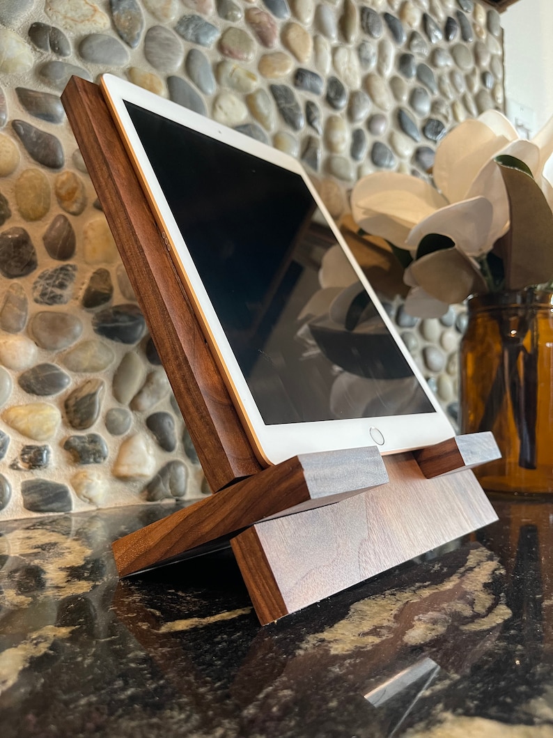 May include: A wooden tablet stand with a dark walnut finish. The stand is angled to hold a tablet upright. The stand is made of two pieces of wood that are joined together.