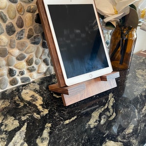May include: A wooden tablet stand with a dark brown finish. The stand is made of solid wood and has a simple, modern design. The stand is designed to hold a tablet in a vertical position, allowing for easy viewing.