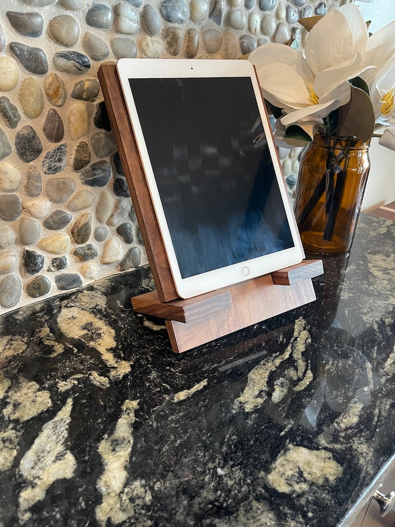 May include: A wooden tablet stand with a dark brown finish. The stand is made of solid wood and has a simple, modern design. The stand is designed to hold a tablet in a vertical position, allowing for easy viewing.
