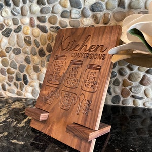 May include: A wooden kitchen conversion chart with a stand. The chart has measurements for gallons, quarts, pints, cups, ounces, and milliliters. The text on the chart reads "Kitchen Conversions".