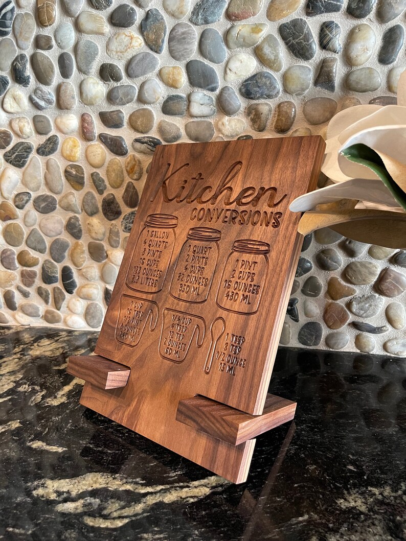 May include: A wooden kitchen conversion chart with a stand. The chart has measurements for gallons, quarts, pints, cups, ounces, and milliliters. The text on the chart reads "Kitchen Conversions".