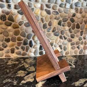 May include: A wooden phone stand with a natural wood finish. The stand is made of two pieces of wood that are joined together at a right angle. The stand is designed to hold a phone in a vertical position.