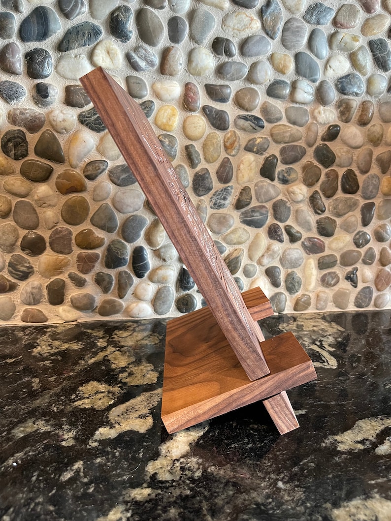 May include: A wooden phone stand with a natural wood finish. The stand is made of two pieces of wood that are joined together at a right angle. The stand is designed to hold a phone in a vertical position.