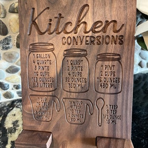 May include: A wooden kitchen conversion chart with engraved measurements for gallons, quarts, pints, cups, ounces, and milliliters. The chart is labeled "Kitchen Conversions".