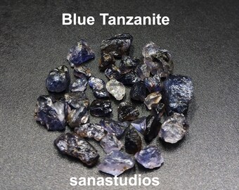 May include: A pile of blue Tanzanite gemstones, rough and unpolished, on a dark gray background. The text "Blue Tanzanite" and "sanastudios" are visible in the image.