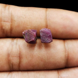 May include: Two rough ruby gemstones, displaying a deep red-purple colour. The stones have a natural, unpolished appearance, with visible texture and colour variations. The gemstones are shown against a neutral background.
