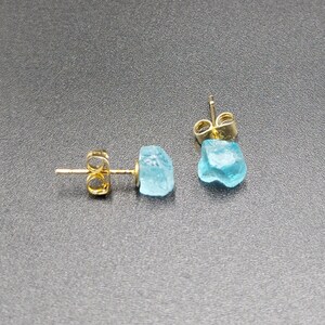 May include: A pair of gold stud earrings with light blue gemstone accents. The gemstones are irregularly shaped and have a rough, natural look.