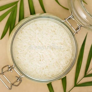 Op de afbeelding: Een heldere glazen pot gevuld met witte rijst, gezien van bovenaf. De pot heeft een metalen sluiting en een heldere glazen deksel. Groene palmbladeren zijn rond de pot gerangschikt op een beige oppervlak.