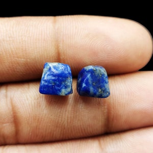 Puede incluir: Dos pequeñas piedras de lapislázuli de corte cuadrado. Las piedras son de color azul intenso con motas doradas y grises. Las piedras son lisas y pulidas, y parecen ser piedras preciosas.
