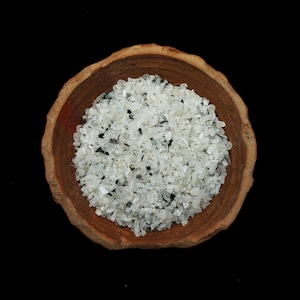 Op de afbeelding: Een houten kom gevuld met kleine, gemalen witte en zwarte stenen. De kom heeft een natuurlijke, rustieke uitstraling met een lichtbruine kleur. De stenen zijn verspreid over de kom en creëren een textuur op een zwarte achtergrond.