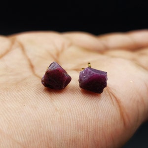 May include: A pair of raw ruby stud earrings. The earrings feature rough-cut, deep red-purple gemstones with gold-colored posts. The earrings are displayed on a light-toned surface.