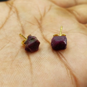 Może przedstawiać: Para kolczyków sztyftów z rubinami i złotymi sztyftami. Surowe, nieoszlifowane rubiny mają głęboki czerwony kolor, a złote sztyfty mają prosty, klasyczny design. Kolczyki są prezentowane na jasnym odcieniu skóry.