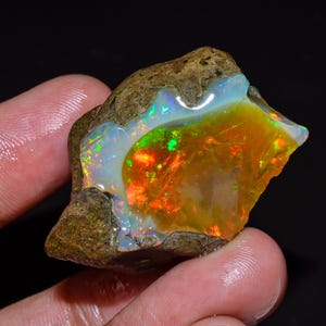 May include: A raw opal gemstone displaying a spectrum of colours. The stone shows orange, green, and blue tones, with a milky white base. The opal is set against a dark backdrop, accentuating its natural beauty.