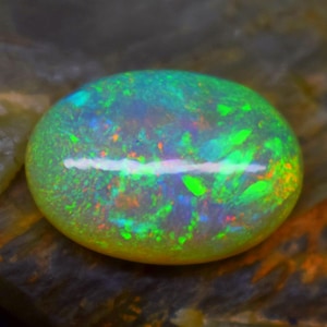 May include: A vibrant, oval-shaped opal gemstone with a mesmerizing play of color. The stone displays a spectrum of greens, blues, and oranges, with a bright, iridescent sheen. The opal is set against a natural, textured background.