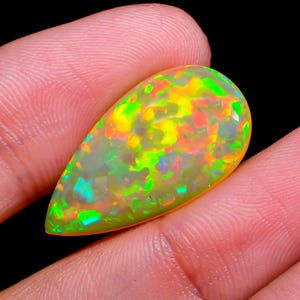 May include: A vibrant, pear-shaped opal gemstone. The stone displays a kaleidoscope of colors, including green, yellow, orange, and hints of blue. The opal is held between fingers against a black background, highlighting its iridescent qualities.