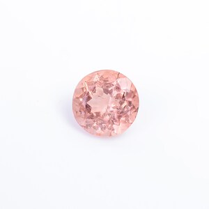 K&ouml;nnte beinhalten: Ein runder, facettierter Edelstein in einem zarten Pfirsichton. Der Edelstein ist vor einem wei&szlig;en Hintergrund platziert, der seine Klarheit und Brillanz hervorhebt. Der Schliff des Steins erzeugt viele Facetten.