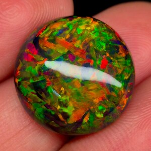 May include: A round, polished opal gemstone with a vibrant display of colors. The opal features a mix of green, orange, red, and blue hues, with a prominent white streak across the center. The gemstone is held in a hand, showcasing its natural beauty.