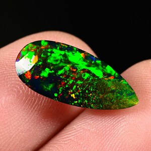 May include: A teardrop-shaped black opal gemstone with vibrant green, red, and orange flashes. The polished surface reflects light, showcasing the stone's internal color play. The opal is held between fingers.