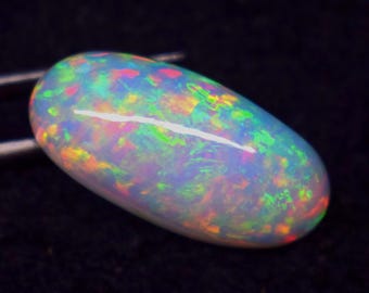May include: A polished, oval-shaped opal gemstone. The opal displays a vibrant play-of-color, with flashes of green, blue, pink, and orange hues across a milky white base. The gemstone is held by a pair of tweezers against a dark background.
