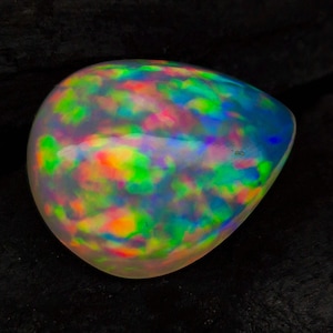 Op de afbeelding: Een gepolijste, ovaalvormige opaal edelsteen. De opaal vertoont een levendig kleurenspel, met flitsen van groen, blauw, geel, oranje en rood. De edelsteen is geplaatst tegen een donkere achtergrond, wat de iriserende kwaliteiten benadrukt.