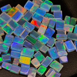 Puede incluir: Una colección de gemas de ópalo rectangulares con colores iridiscentes, incluyendo azul, verde, amarillo y rojo. Los ópalos están dispuestos muy juntos, mostrando sus patrones y texturas únicos. Las gemas se encuentran sobre un fondo oscuro.