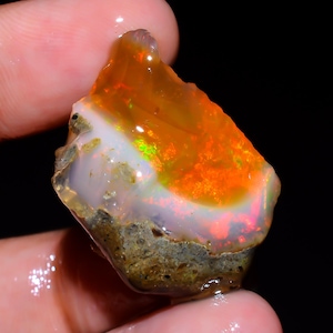 May include: A rough-cut opal gemstone with vibrant orange, white, and green hues. The stone has a layered appearance with a rough, textured base. The opal is held in a hand, showcasing its natural beauty.