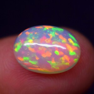 Può includere: Primo piano di una vibrante pietra preziosa di opale ovale. L'opale mostra un colore di base bianco latte con lampi di verde, arancione e giallo, creando un effetto di gioco di colori. La gemma è tenuta tra le dita su uno sfondo scuro.